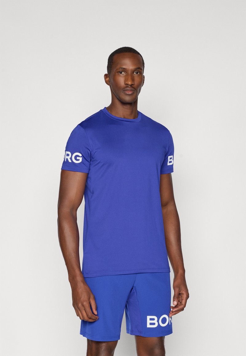 Ανδρική αθλητική εμφάνιση με ένα μπλε μονόχρωμο κοντομάνικο πουκάμισο και αντίστοιχες βερμούδες, και τα δύο με λευκό κείμενο "BORG" στα μανίκια και τις βερμούδες.