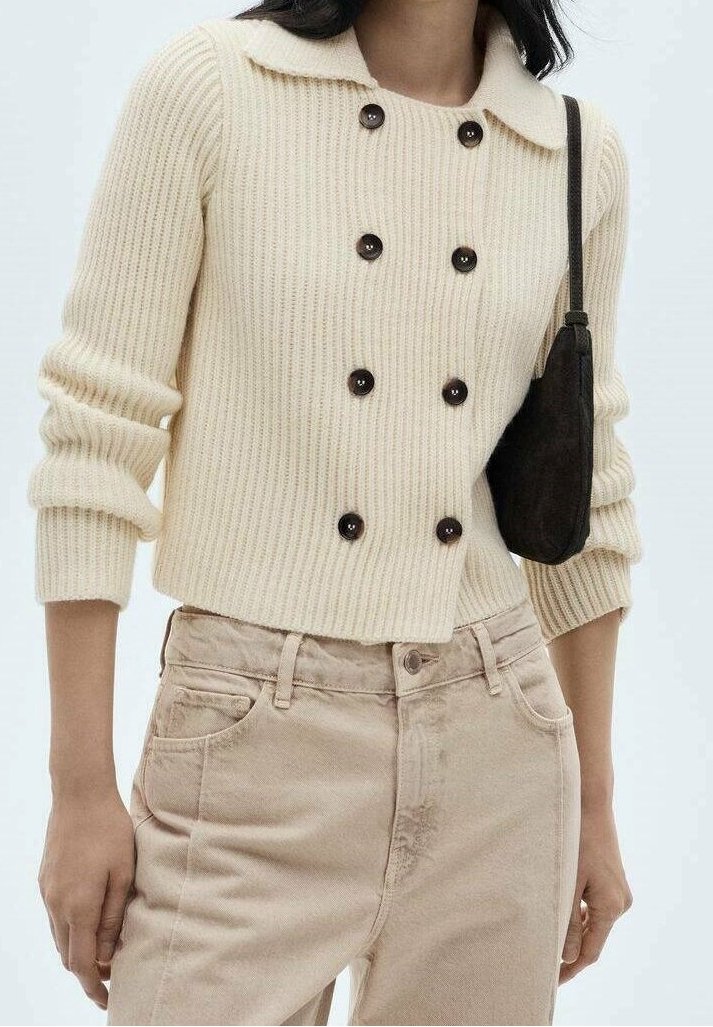 Femme portant un cardigan en maille côtelée crème avec des boutons noirs, un pantalon beige taille haute et un petit sac à bandoulière noir.