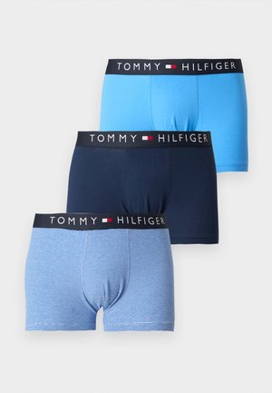 Homme musclé debout pieds nus sur fond blanc, portant un boxer bleu marine Tommy Hilfiger avec une ceinture à logo.