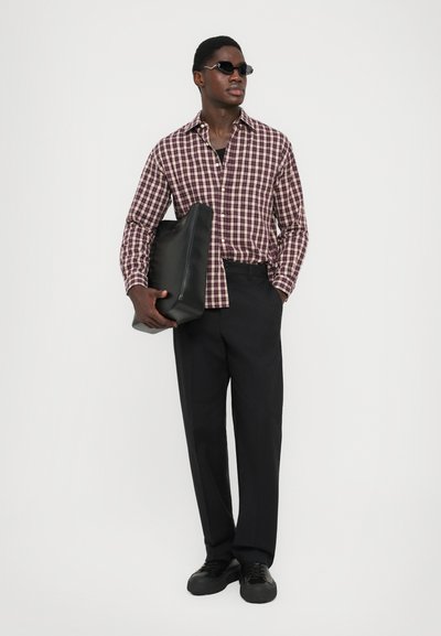 Uomo con occhiali da sole, camicia a quadri bordeaux, pantaloni neri, che tiene una grande borsa di pelle nera contro uno sfondo chiaro e semplice.