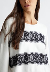 Weißer Pullover mit zwei horizontalen schwarzen Spitzenbändern mit fransigen Kanten. Der Stoff hat eine strukturierte Oberfläche und eine lockere Passform.
