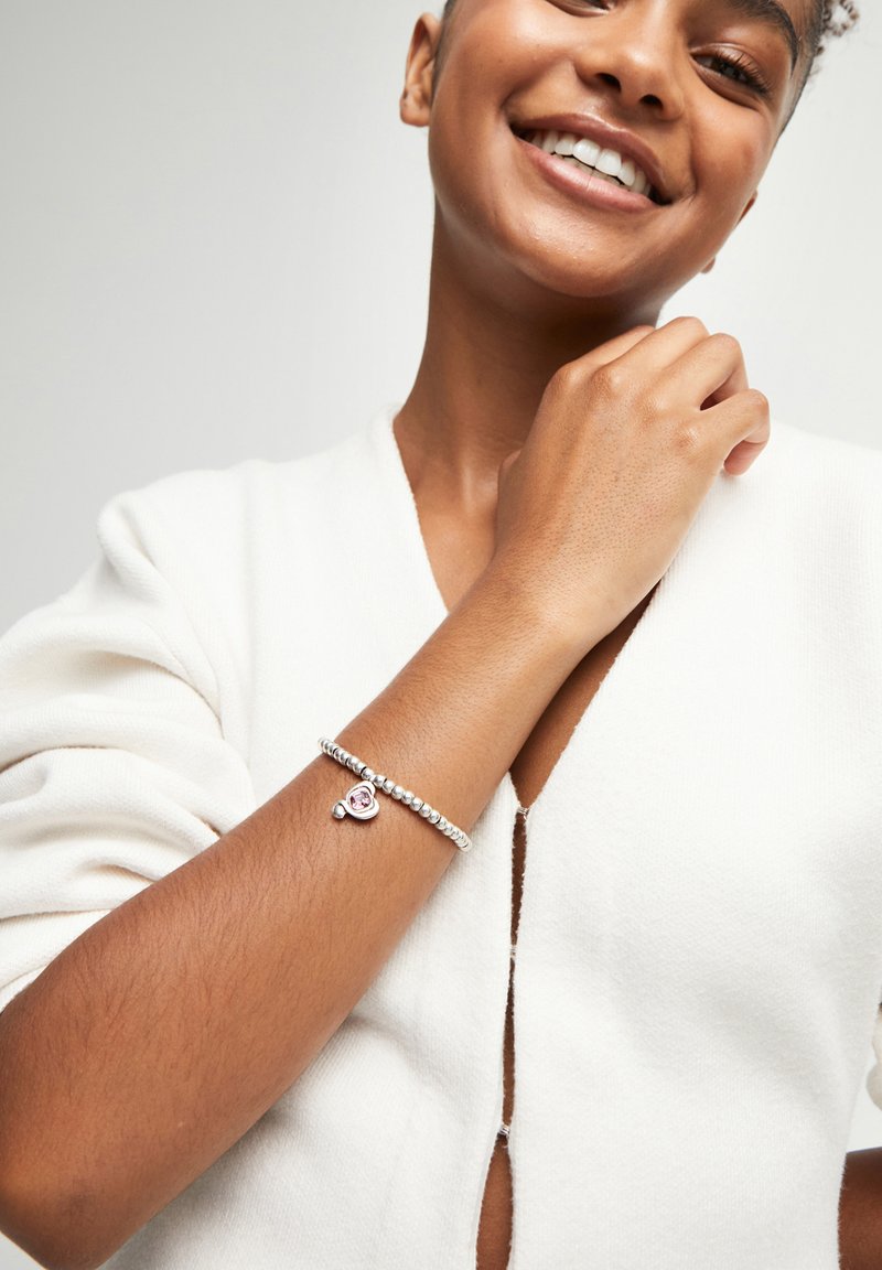 Une personne souriante en haut blanc montre un bracelet en perles argentées avec un charme en forme de cœur rose sur le poignet gauche, sur fond uni.