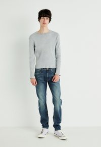 Szary prążkowany T-shirt z długim rękawem, wyblakłe niebieskie dżinsy i białe sneakersy. Model pozuje na jednolitym białym tle.