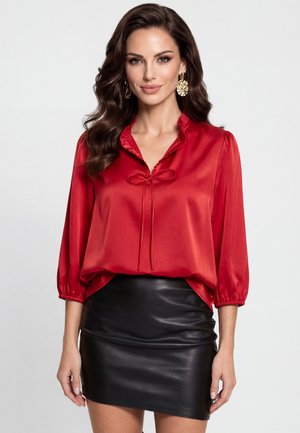 Vrouw met lang golvend bruin haar die gouden oorbellen draagt, een rode satijnen blouse met driekwart mouwen en een zwarte leren minirok.