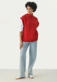 Top roșu din tricot fără mâneci, cu guler înalt, purtat cu blugi largi ușor albaștri și adidași albi. Păr creț, accesorii minime.