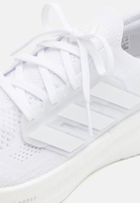 Primo piano di una sneaker in tessuto lavorato a maglia bianco con lacci bianchi annodati e tre strisce bianche sul lato, con suola ammortizzata.