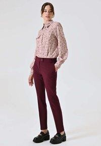Blusa rosa chiaro con motivo floreale, abbinata a pantaloni color bordeaux. Mocassini neri con suole spesse e dettagli dorati completano il look.