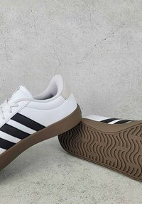 Biele športové tenisky s čiernymi pruhmi, textúrovaný syntetický zvršok, polstrovaná manžeta a hnedá gumená podrážka s vlnkovým vzorom.