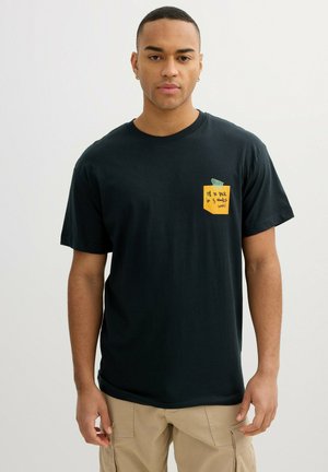 Joven con una camiseta negra con un gráfico de una nota adhesiva amarilla que dice "Volveré en 5 minutos (pronto)" en el pecho, pantalones beige.