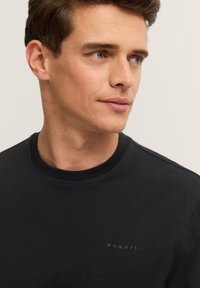 Junger Mann mit kurzen braunen Haaren, der ein schwarzes Rundhals-Shirt mit "Bugatti"-Logo trägt und leicht nach rechts schaut, vor einem schlichten Hintergrund.