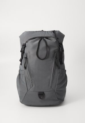 Mochila gris de material de lona, que cuenta con correas ajustables, dos cremalleras delanteras, bolsillos laterales y un cierre con cordón en la parte superior.