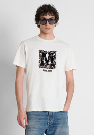 Uomo con occhiali da sole quadrati neri, t-shirt bianca con lettera M floreale nera e scritta Morato, e jeans blu, in piedi con una mano in tasca.