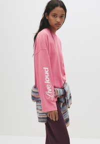 Tânără îmbrăcată cu un tricou roz cu mâneci lungi pe care scrie "Trăiește tare" pe mânecă, pulover tricotat cu dungi legat la talie și pantaloni închiși la culoare.