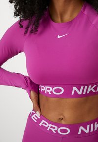 Dugih rukava, pripijen majica za sport u vibrantnoj ružičastoj boji s bijelim Nike logotipom; sadrži odgovarajući elastični pojas s "NIKE PRO" tekstom.