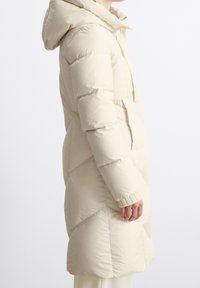 Vaaleanbeige puffatakki hupulla, jossa on tilkkutöistämätön muotoilu, teksturoitu kangas ja ribbireunat. Polvenpituinen, vetoketjukiinnityksellä ja etutaskulla.