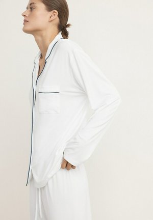 Femme aux cheveux retenus, portant un haut de pyjama blanc à manches longues avec des liserés noirs et un pantalon de pyjama blanc assorti, debout de profil.