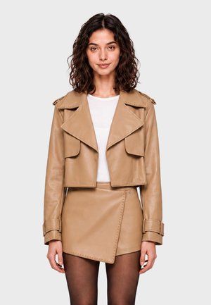 Femme aux cheveux bouclés foncés porte une veste en cuir courte couleur fauve, une jupe en cuir assortie, un haut blanc et des collants noirs transparents, debout devant un fond blanc.
