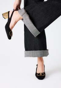 Zapatos de ante negro con detalles decorativos en dorado y un tacón cuadrado texturizado, combinados con pantalones de mezclilla oscuros vueltas.