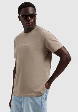 Man draagt een beige t-shirt met de tekst "Pure Path", lichtblauwe spijkerbroek en donkere zonnebril, staand met één hand in de zak.