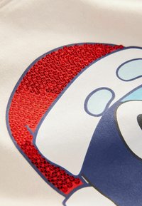 Gros plan sur un ours cartoon bleu et blanc portant un chapeau de Père Noël rouge texturé et brillant sur un fond de tissu crème.