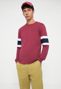 Burgundi pikkade varrukatega särk, millel on valged ja tumesinised triibud varrukatel, ümmargune kaelus ja väike GAP logo. Kandmine kollaste pükstega.