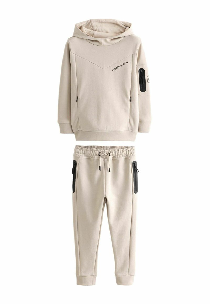 Beige urheiluvaatteet, jotka koostuvat hupparista, jossa on vinosaumat ja sivuilla vetoketjulliset taskut, sekä joustovyötäröisistä housuista, joissa on myös vetoketjulliset taskut.