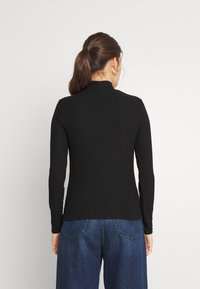 Top lungo a maniche lunghe nero a costine con colletto alto, caratterizzato da una vestibilità aderente e una superficie testurizzata, indossato con jeans blu larghi.