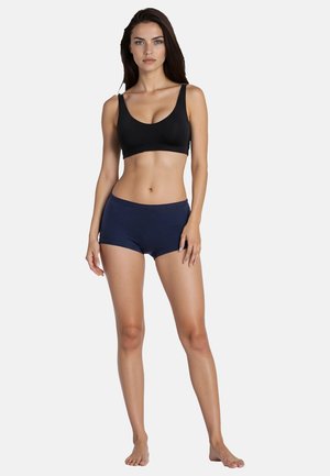 Frau steht barfuß da, trägt einen schwarzen Sport-BH und marineblaue Boyshorts vor einem schlichten weißen Hintergrund.