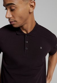 Donkerpaarse polo met een structuurstof, voorzien van een klassieke kraag, een drieknopenlijst en een klein logo op de borst.