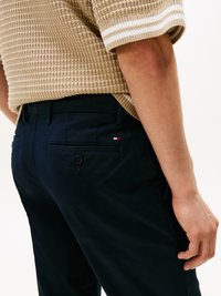 Navyblå bukser med en glat overflade, der har en baglomme med knap og et lille mærke med logo. Båret med en beige striktrøje.