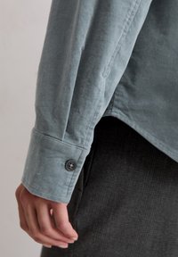 Langärmeliges Hemd aus weichem, hellblauem Stoff mit Knopfmanschetten und lockerem Schnitt, kombiniert mit einem strukturierten, dunklen Stoff darunter.