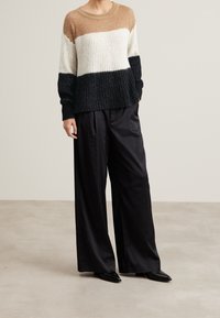 Pull en tricot avec trois blocs de couleur : beige, blanc et noir. Pantalons large noirs en tissu lisse. Manches longues et col rond.