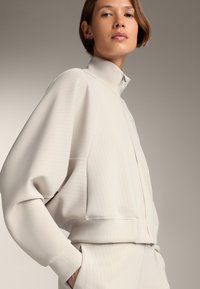 Vaaleanharmaa korkeakauluksinen takki, jossa on pystysuorat raidat, rento malli, vetoketjusuljenta ja ribbauseristicat. Silkkinen pinta, minimalistinen muotoilu.