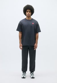 Mørkegrå bomulds-t-shirt med et lille rødt logo, parret med matchende grå bukser og hvide atletik-sko. Modellen står foran en lys baggrund.