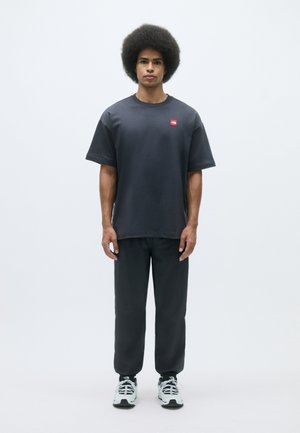 Σκοτεινό γκρι βαμβακερό T-shirt με ένα μικρό κόκκινο λογότυπο, συνδυασμένο με αντίστοιχες γκρι παντελόνες και άσπρα αθλητικά παπούτσια. Το μοντέλο στέκεται σε ένα ανοιχτό φόντο.