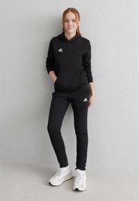 Ung person som står mot en grå vägg iförd en svart Adidas hoodie och byxor med vita sneakers, med händerna i fickorna och ett avslappnat uttryck.