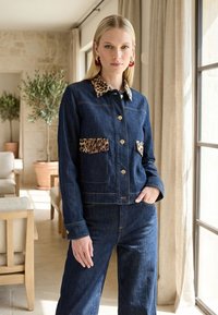 Femme portant une veste en denim foncé et un pantalon avec un col et des rabats de poche à imprimé léopard, se tenant près d'une fenêtre dans une pièce lumineuse.