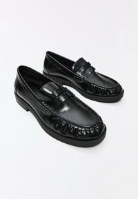 Paire de mocassins penny en cuir noir brillant avec bouts arrondis et petits talons sur fond blanc uni.