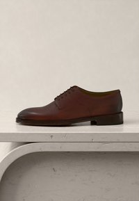 Chaussure Oxford en cuir marron à lacets sur une surface en marbre clair, vue latérale, sur fond beige uni.