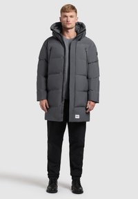 Muškarac u sivom zimskoj jakni s kapuljačom, tamnim hlačama i crnim čizmama, stoji protiv jednostavne svijetle pozadine.