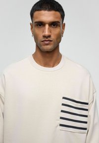 Beige sweatshirt met een oversized pasvorm, voorzien van een gestreepte zwart-wit zak aan de linkerzijde. Zachte stof en ronde halslijn.