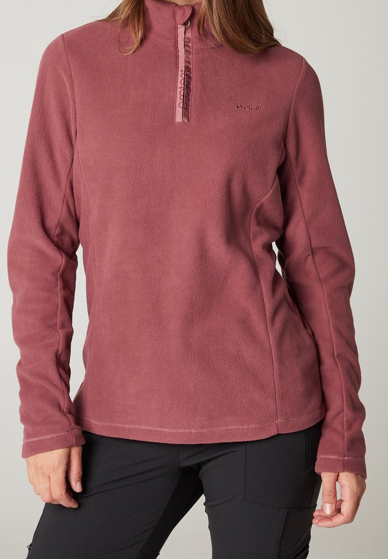 Femme portant un pull polaire rose poudré à demi-fermeture éclair et un pantalon noir, debout les bras détendus sur les côtés.