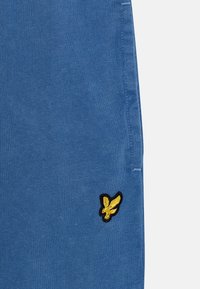 Niebieskie bawełniane szorty z haftowanym żółtym logo ptaka po lewej stronie. Materiał ma gładką teksturę z delikatnym połyskiem.