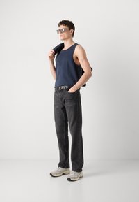 Marine tank topp, mørkegrå jeans og lyse sneakers. Modellen holder en jakke, har solbriller på; uformell passform med rene linjer.