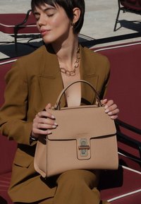 Beige, gestructureerde leren handtas met een textuur, gouden hardware en een sluiting aan de voorkant. Voorzien van een bovenhandvat voor het dragen.