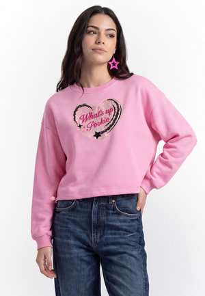 Donna che indossa una felpa rosa con un cuore e il testo "What's up Pookie", abbinata a jeans blu e orecchini rosa a forma di stella.