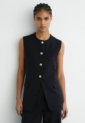 Donna con capelli intrecciati che indossa un gilet nero senza maniche con bottoni dorati, in piedi contro un muro bianco e strutturato.