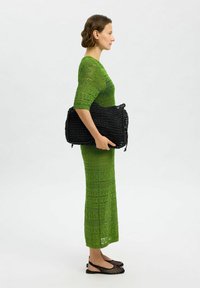 Bolso negro de tejido sostenido contra el perfil lateral de una mujer con un vestido largo, ajustado, de encaje verde y zapatos planos de malla negros.