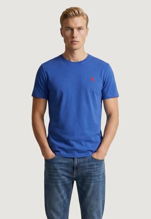 Uomo biondo in piedi con le mani nelle tasche, indossa una T-shirt blu a maniche corte con un logo rosso e jeans blu su uno sfondo semplice.