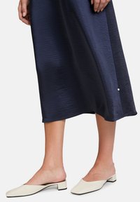 Untere Beine, die einen marineblauen Midi-Rock und off-white, niedrig abgesatzte Slipper mit quadratischen Zehen auf einem weißen Hintergrund tragen.